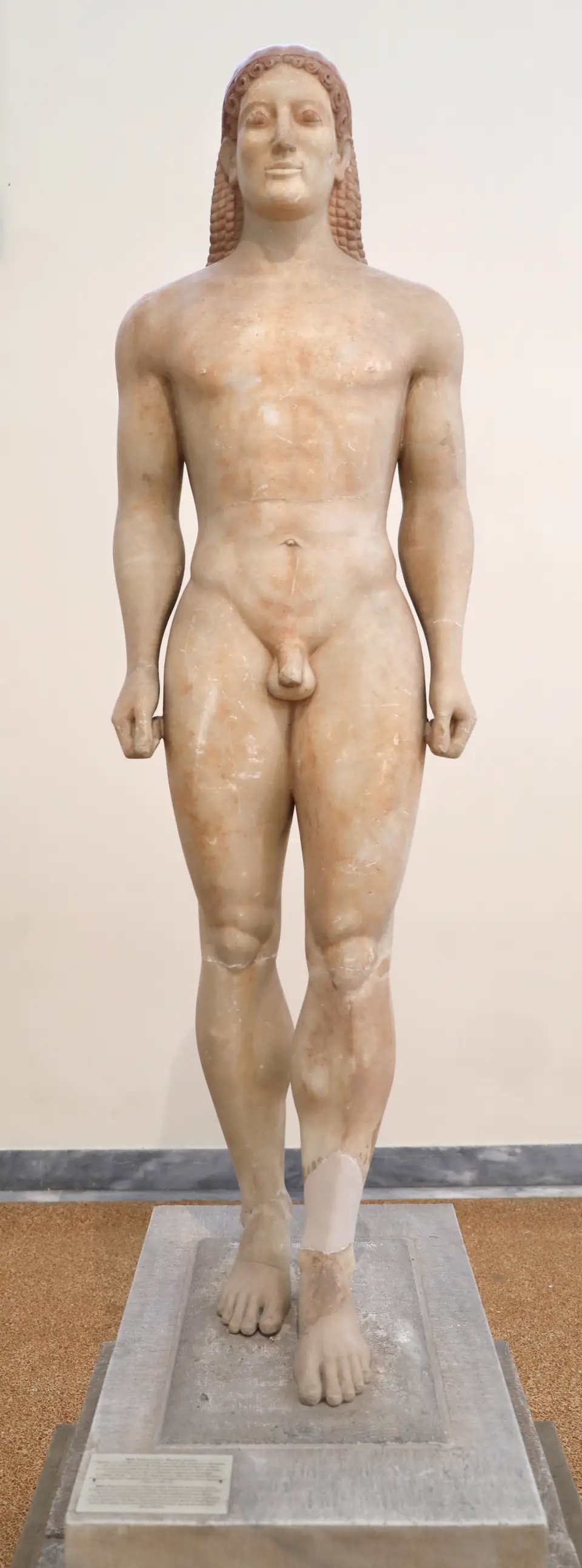 A marble statue of a nude young man standing stiffly with one foot forward, arms at his sides and a slight smile.