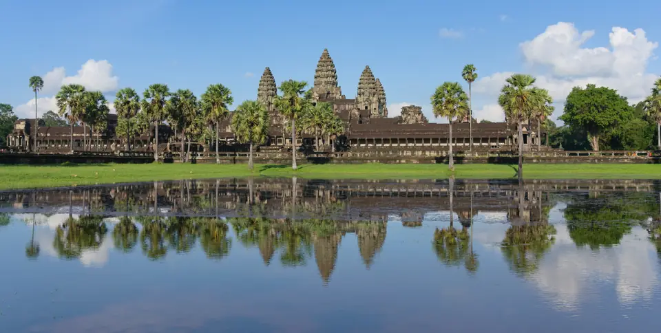 A long stone temple with tall lotus-bud towers reflected in a still pool, with palm trees on either side.