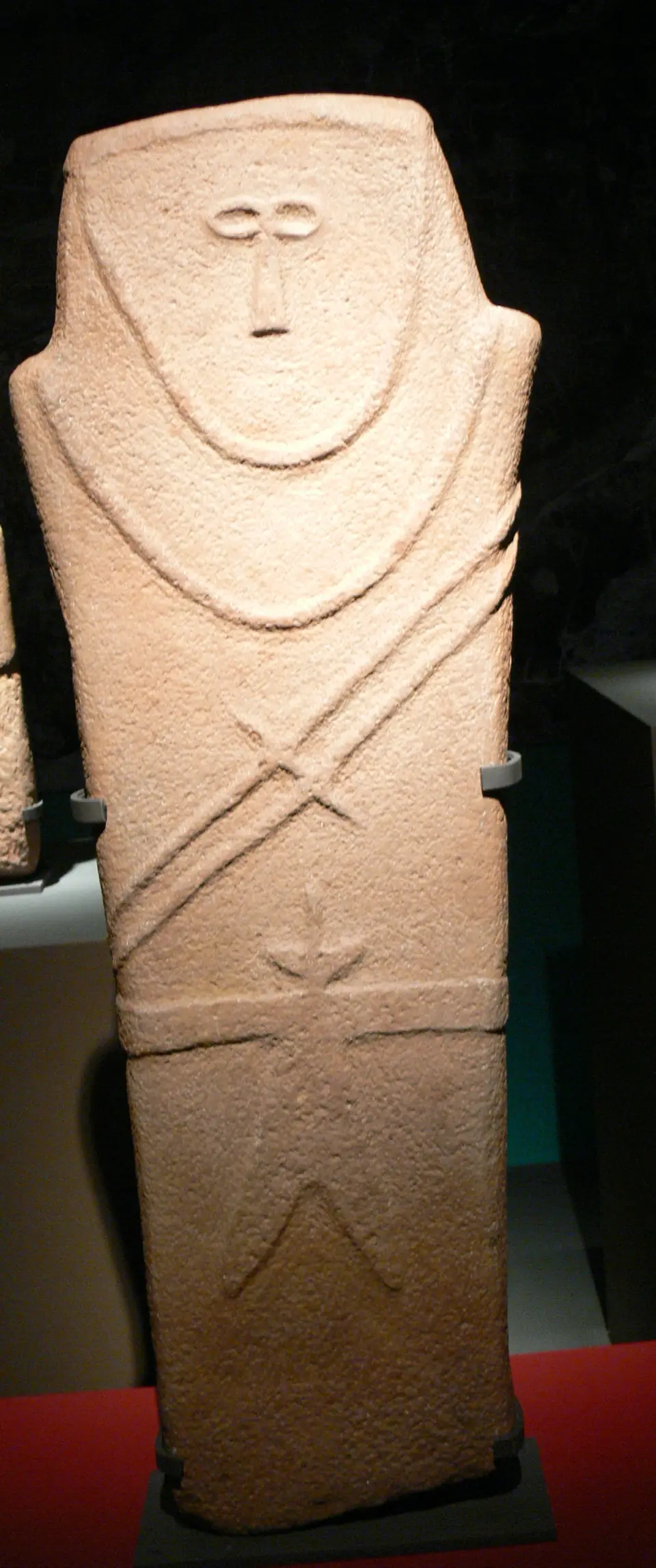 An upright slab of stone carved with a simple human face, arms and a belt in low relief.