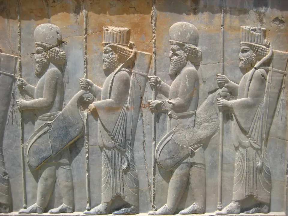 A stone stairway wall carved in low relief with a row of bearded figures holding spears, walking in procession, some in tall fluted hats and some in rounded caps.
