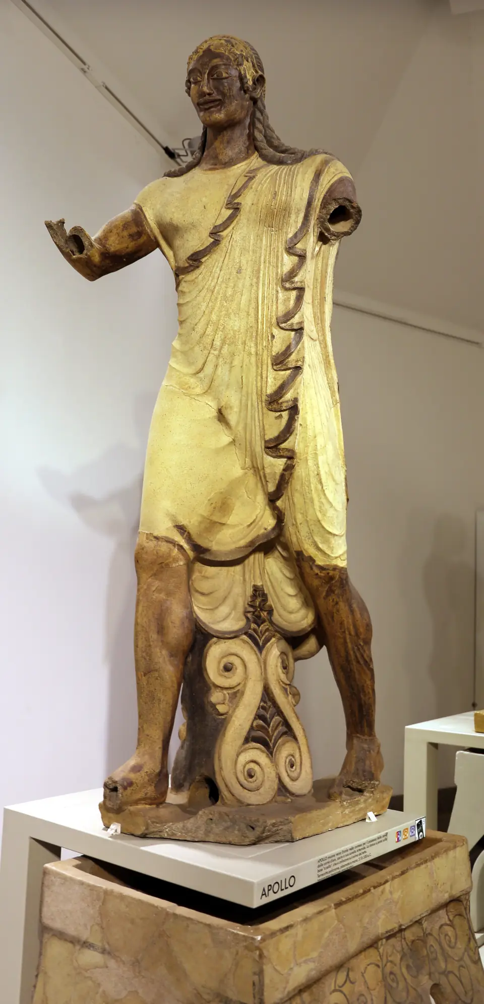 A painted terracotta statue of a striding male figure with a wide smile, wearing a patterned robe.