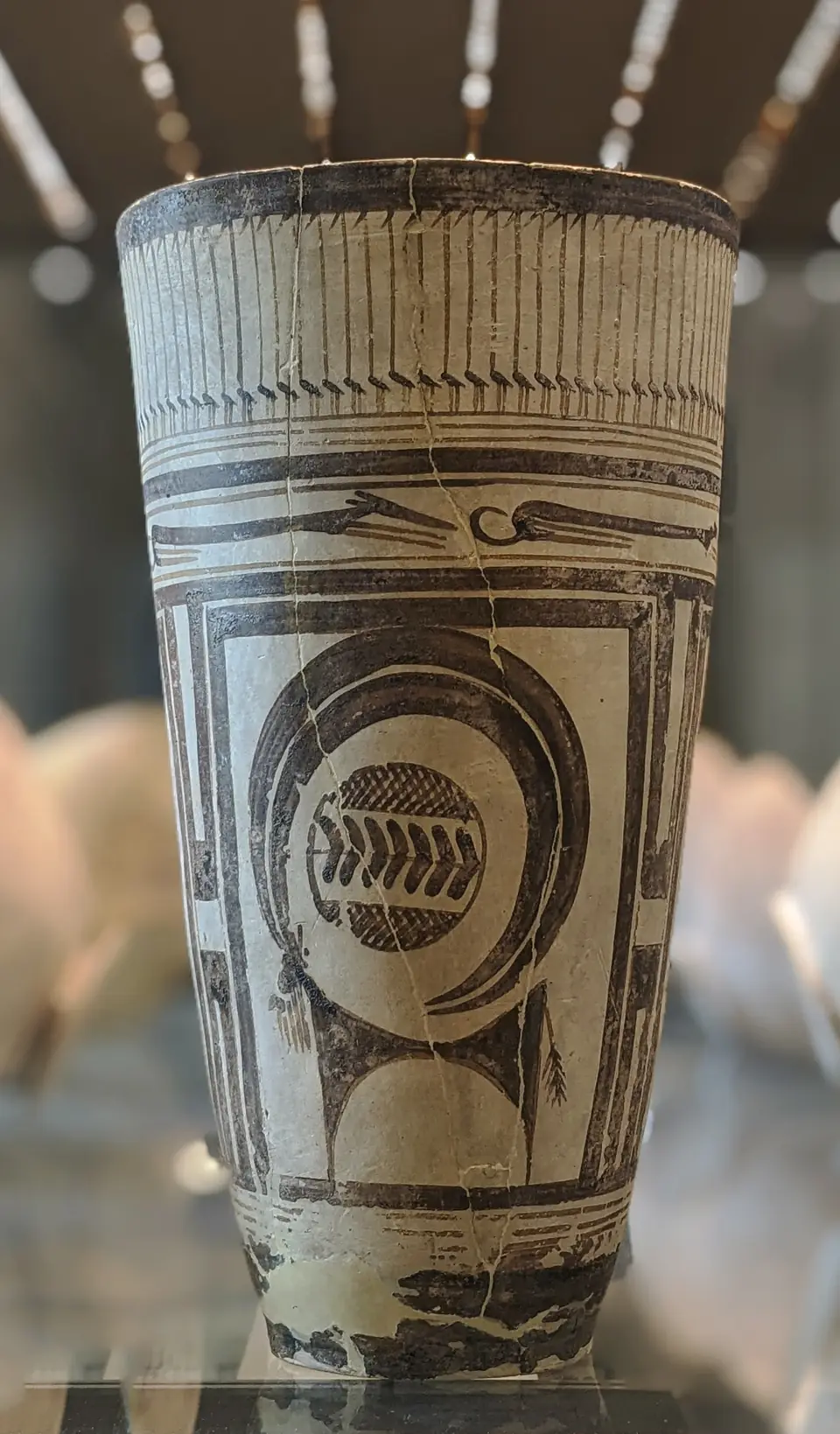 A tall, thin-walled pale clay beaker painted in dark brown with a stylized horned animal inside a panel, long-necked birds along the rim and running dogs in a band.