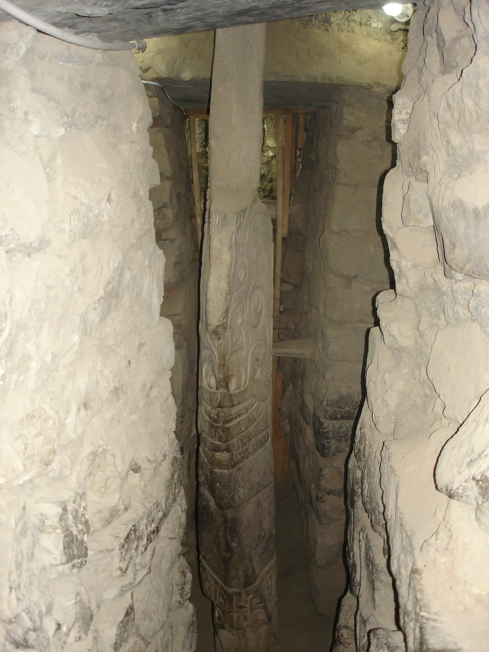 A tall, blade-shaped granite stone in a narrow stone passage, carved with a fanged figure.