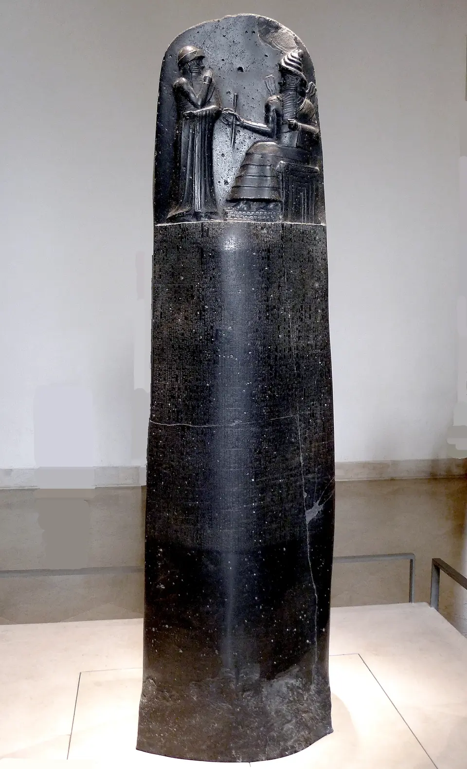 A tall black stone pillar with a relief of a standing ruler before a seated god at the top, and rows of cuneiform writing below.