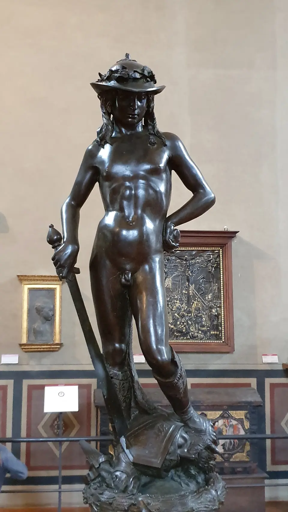 A bronze statue of a slender nude youth in a brimmed hat and boots, one foot on a severed head, holding a sword.
