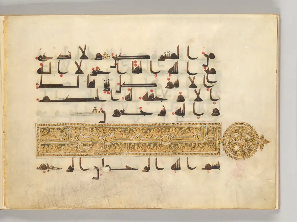 A wide parchment page with five lines of bold, horizontal brown script dotted in red, above a gold decorated band with a round gold ornament in the margin and one more line of script below.