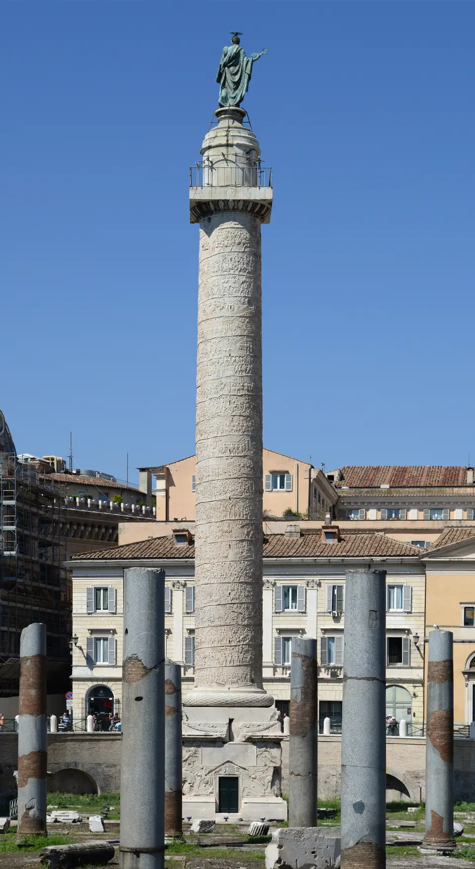 A tall marble column wrapped in a spiral band of carved reliefs and topped by a bronze statue rises behind a row of broken granite column shafts, against a deep blue sky.