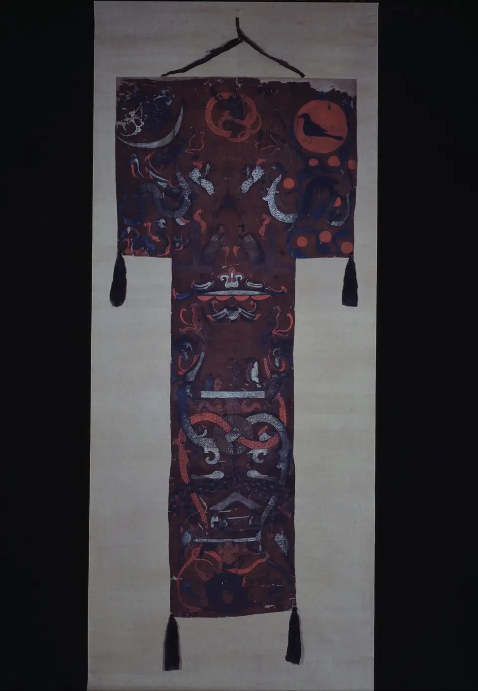 A long T-shaped silk banner on a dark red-brown ground, painted with a crescent moon and a red sun holding a black bird at the top, coiling dragons and figures below, and black tassels hanging from its corners.