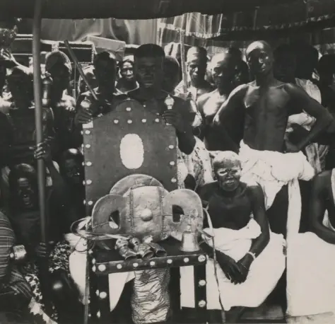 A crowd of people gathered around a gold-covered stool resting on its own chair, in an old black-and-white photograph.
