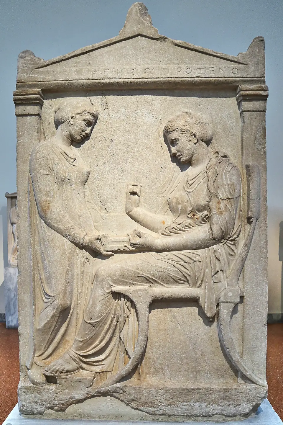 A marble relief of a seated woman lifting an item from a box held by a standing servant, framed like a small temple front.