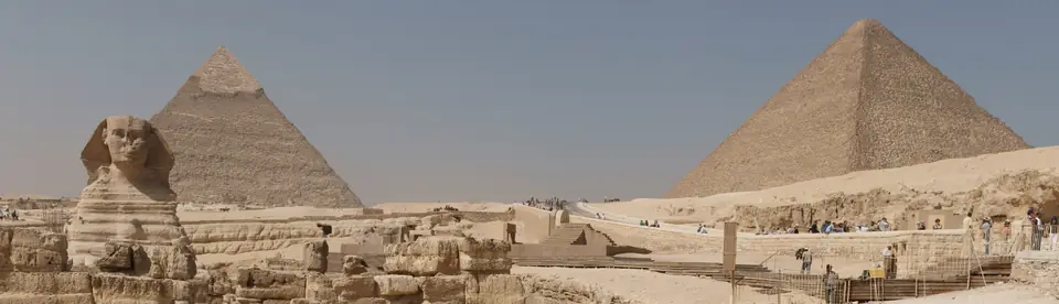 A huge stone sphinx with a human head lies in front of large pyramids in the desert.