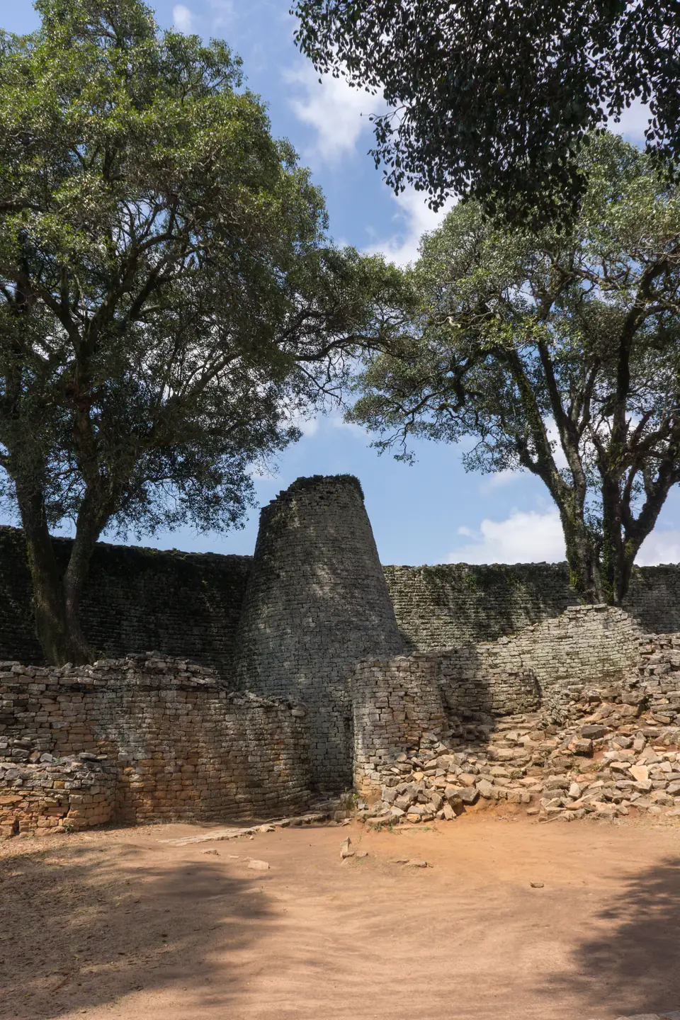 A tall conical stone tower inside a curved wall built of fitted granite blocks, with trees around.