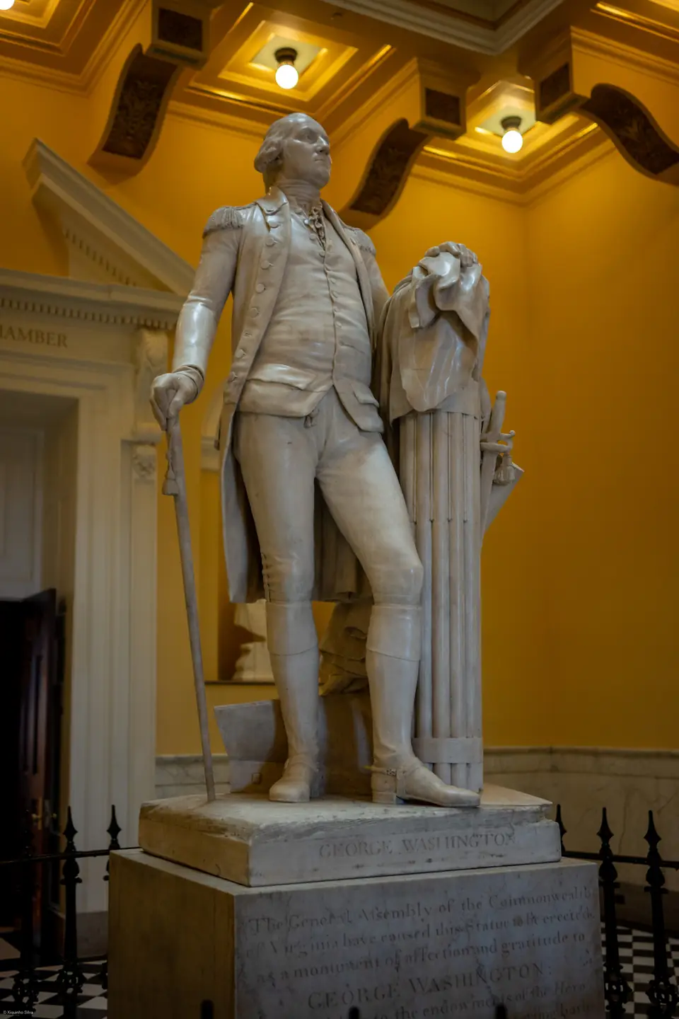 A life-size white marble statue of a man in an 18th-century military coat and boots, one hand on a walking cane and the other resting on a bundle of rods draped with a cloak, in a yellow-walled domed hall.