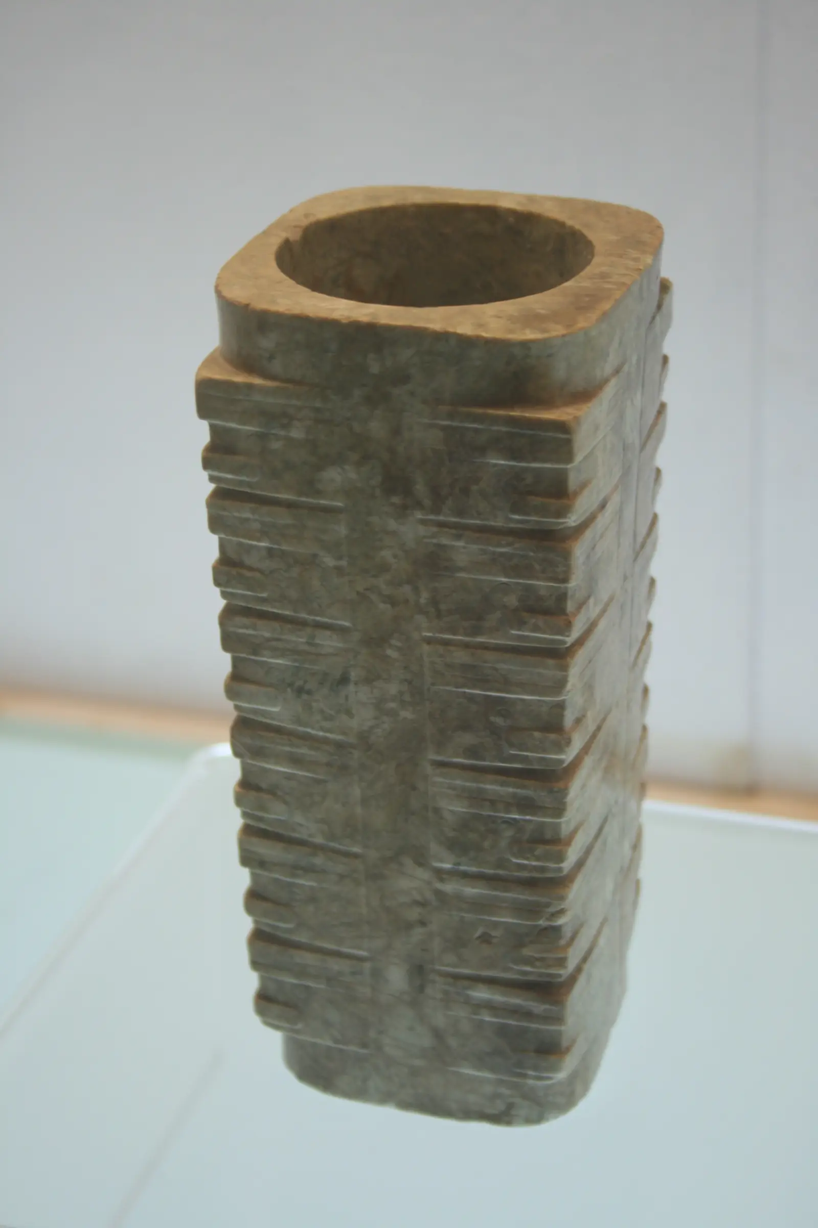 A tall, square jade tube with a round opening at the top, its sides cut into stacked horizontal bands.