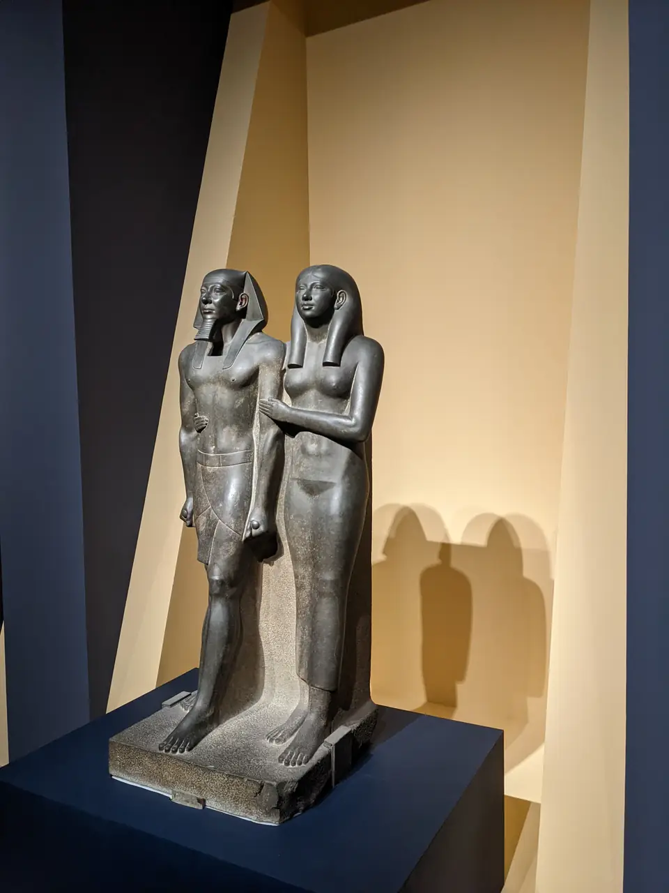 A dark stone statue of a striding king and a queen who embraces him, both standing stiffly and facing forward.