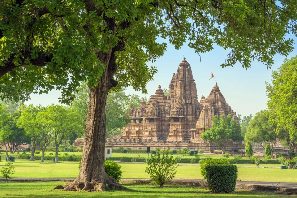 A stone temple with a cluster of tall, rounded, richly carved towers rising from a raised platform, seen behind a large tree.