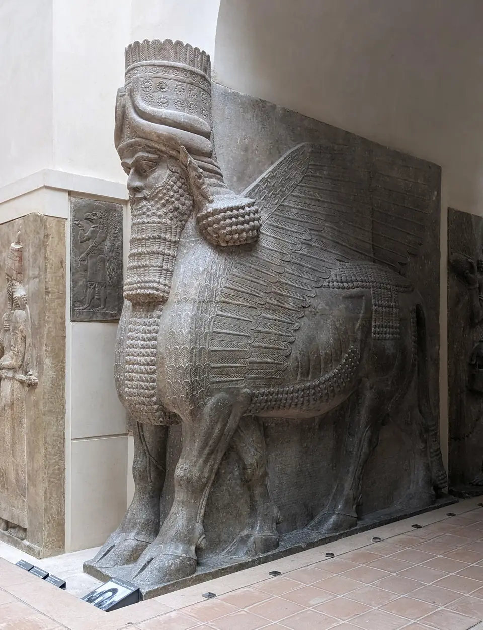 A colossal stone guardian with the body of a bull, feathered wings, a bearded human head and a tall horned crown, set in a doorway.
