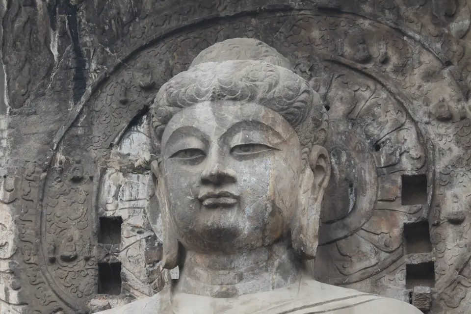 The calm face of a colossal stone buddha carved into a cliff, framed by a carved halo and smaller niches.
