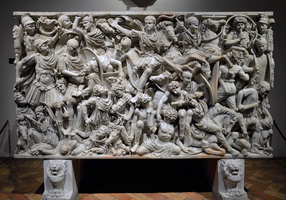A marble coffin carved in deep relief with a dense, chaotic battle, a calm bareheaded rider at the center.