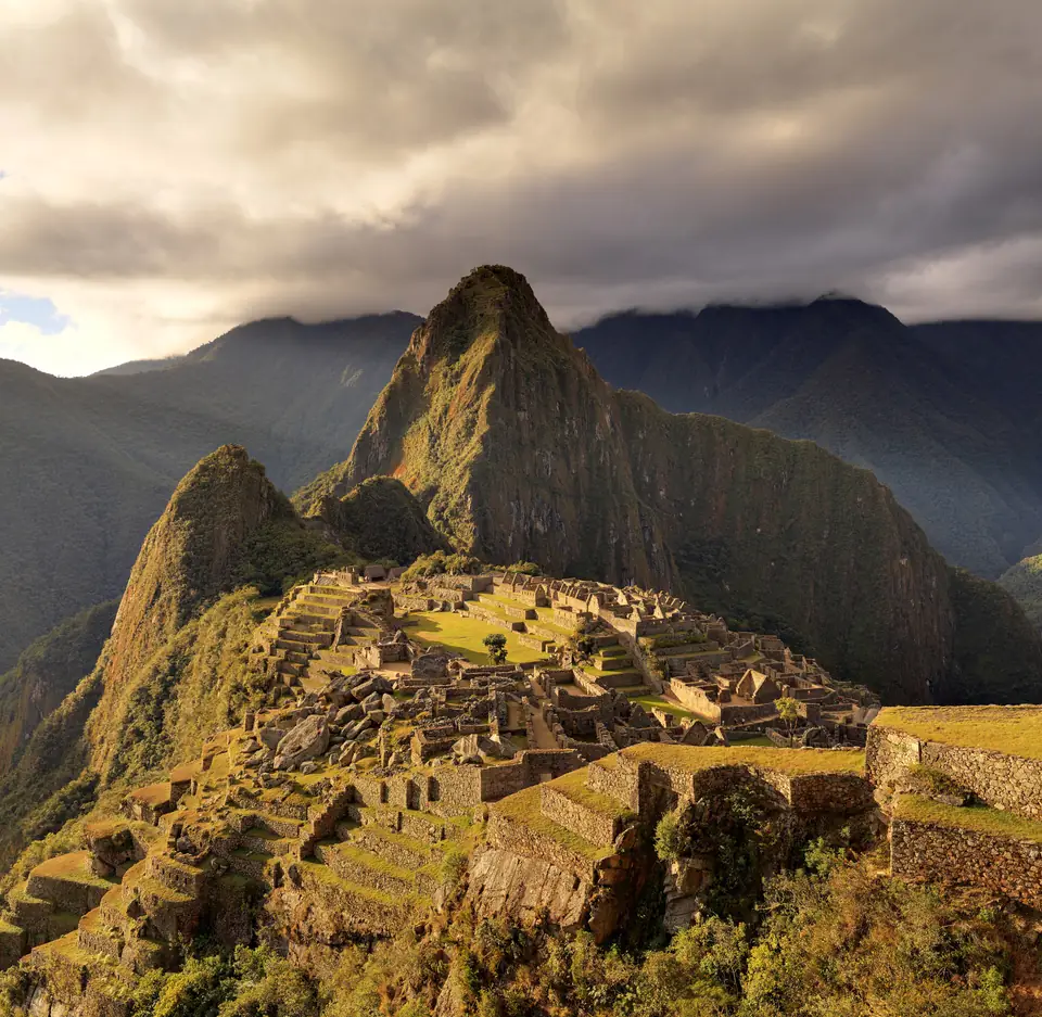 Granite buildings and terraces spread across a high mountain ridge, with steep green peaks behind.