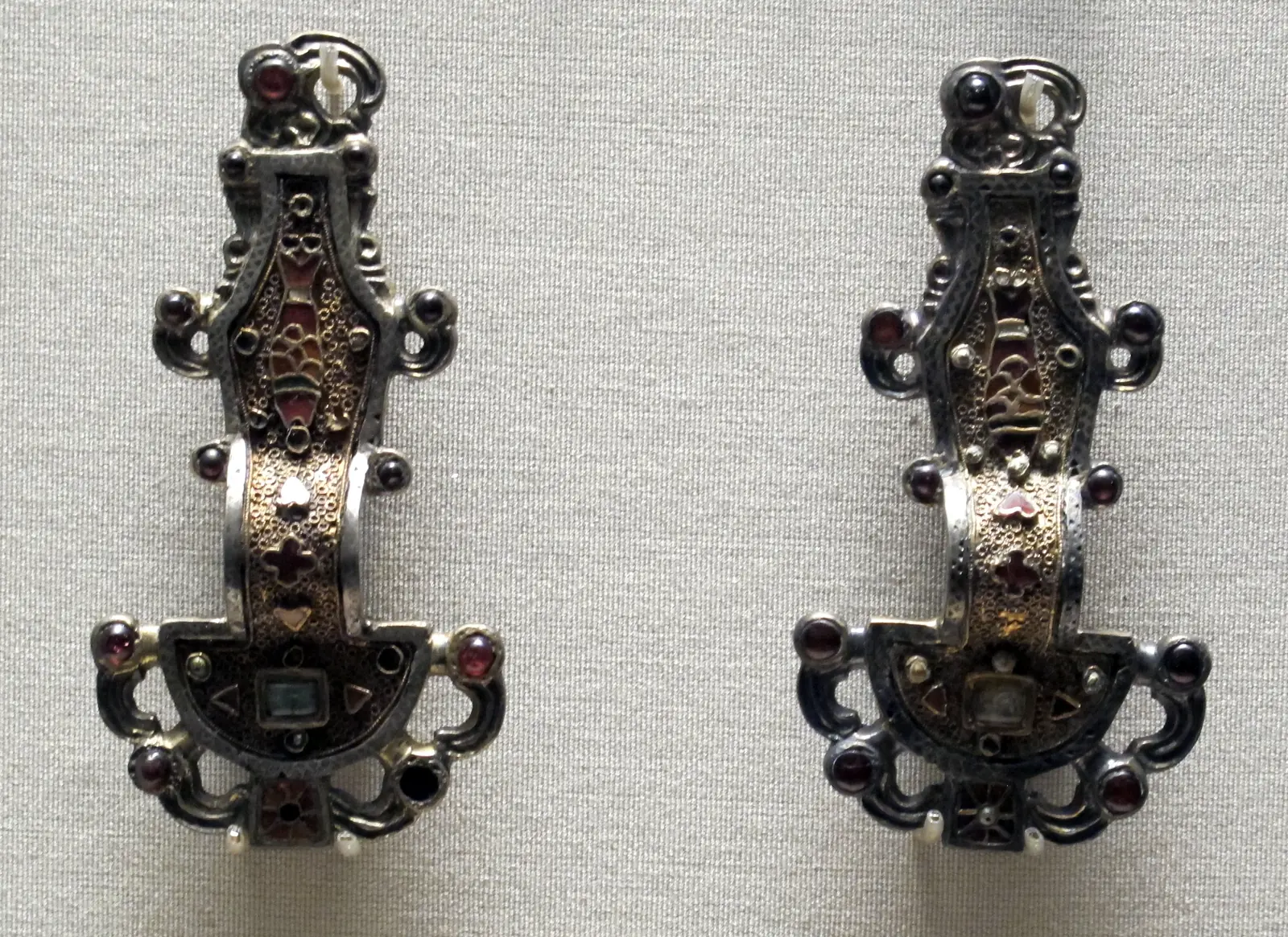 Two matching silver-gilt brooches standing side by side. Each has a half-round plate at the bottom, an arched bow and a long, narrow upper plate inlaid with a fish made of small red garnets; curling hooks with round garnet eyes, like bird heads, project from the edges.