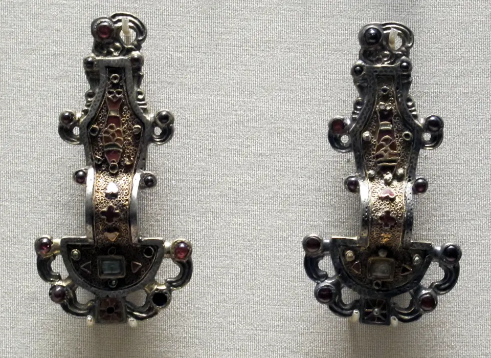 Two matching silver-gilt brooches standing side by side. Each has a half-round plate at the bottom, an arched bow and a long, narrow upper plate inlaid with a fish made of small red garnets; curling hooks with round garnet eyes, like bird heads, project from the edges.