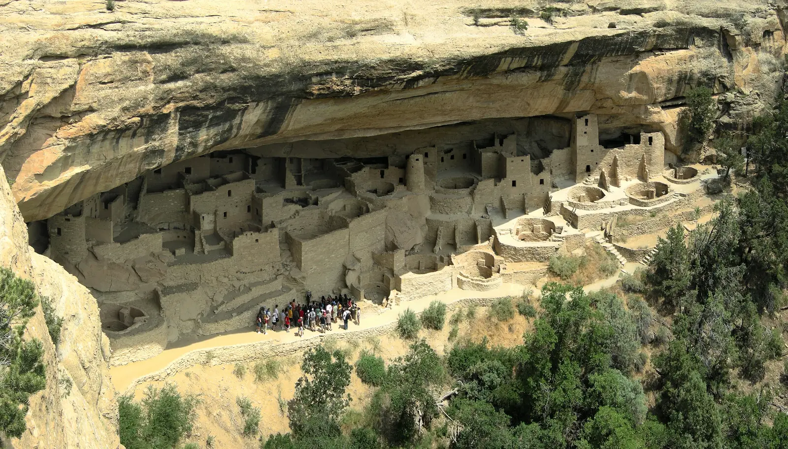Multistory stone buildings and round towers built into a large cave beneath a cliff overhang.
