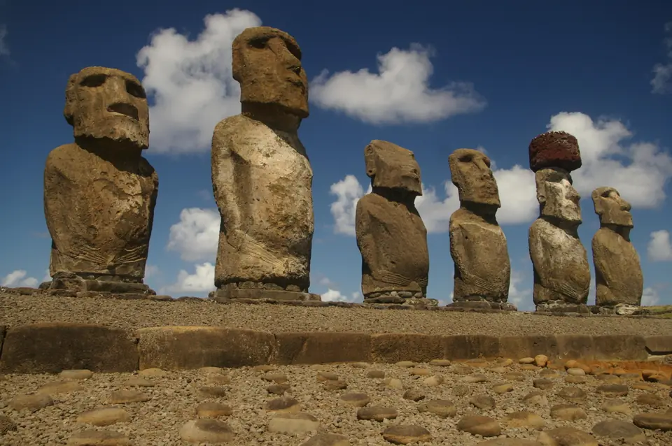 A row of huge stone heads and torsos standing on a stone platform, one wearing a red stone topknot.