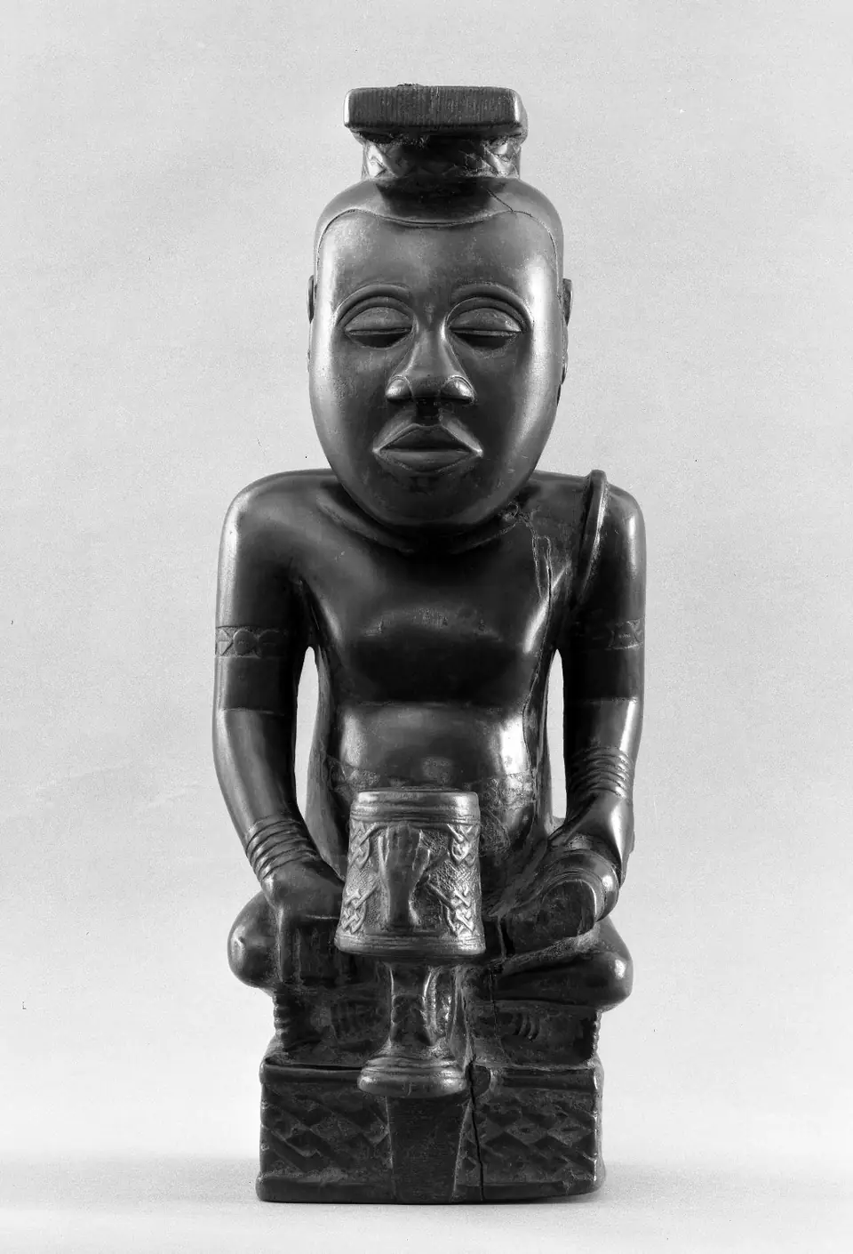 A wooden figure of a seated king with crossed legs, wearing a flat-brimmed headdress, with a small object on the base in front of him.