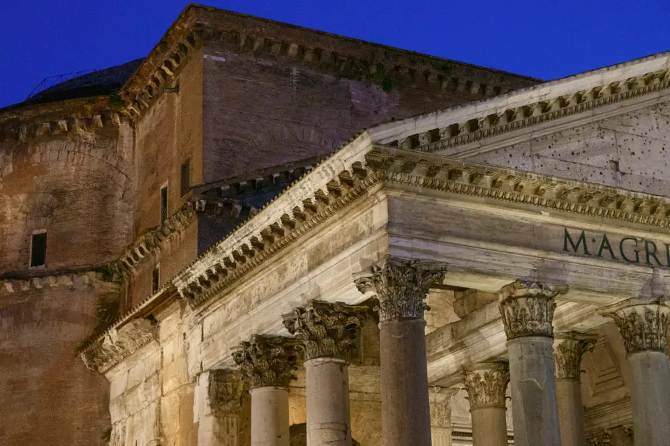 A temple porch of tall columns with leafy capitals under a triangular gable, with an inscription across the front, attached to a massive round brick wall.