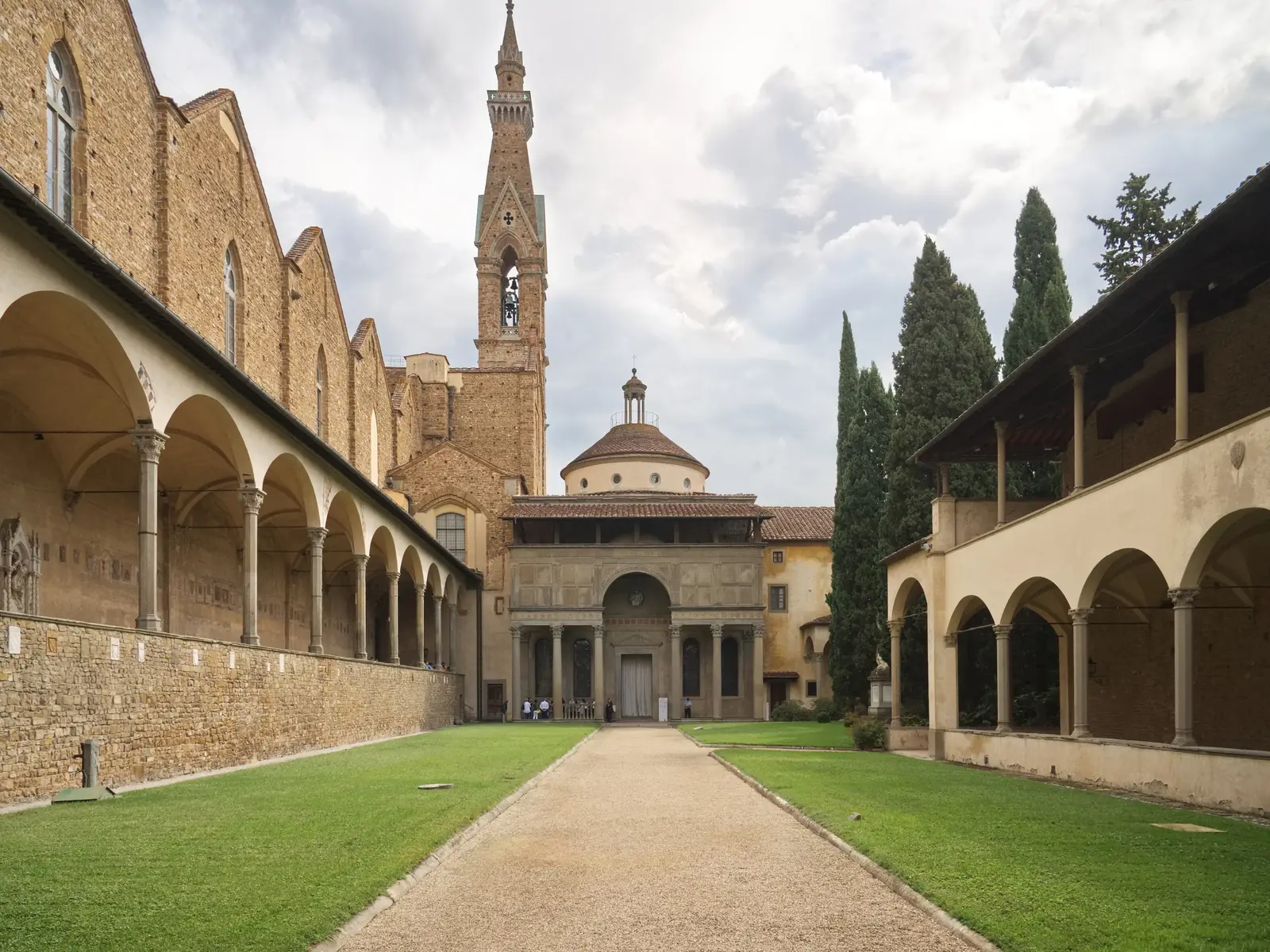 A small Renaissance building with a porch of slender columns, a wide upper wall and a small dome, at the end of a long arcaded courtyard.