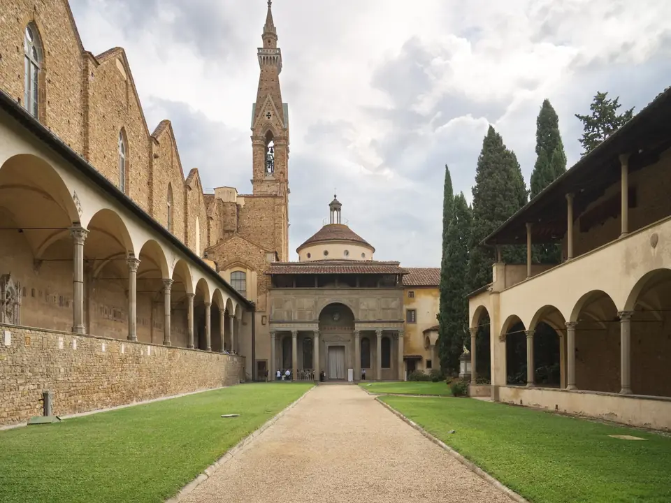 A small Renaissance building with a porch of slender columns, a wide upper wall and a small dome, at the end of a long arcaded courtyard.