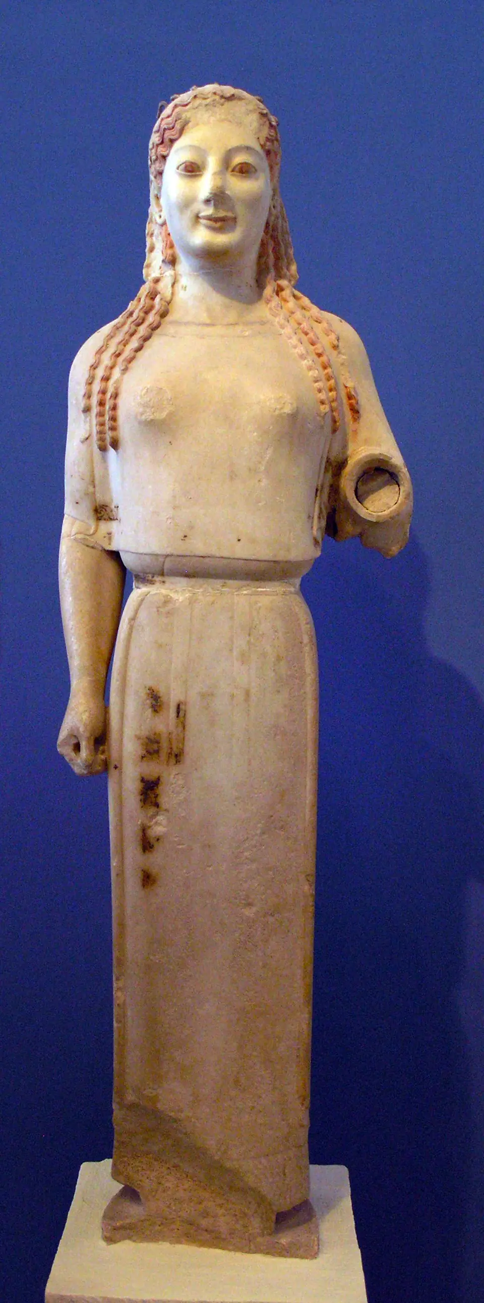 A marble statue of a standing young woman in a straight, belted dress, with long braided hair and a slight smile; one forearm is missing.