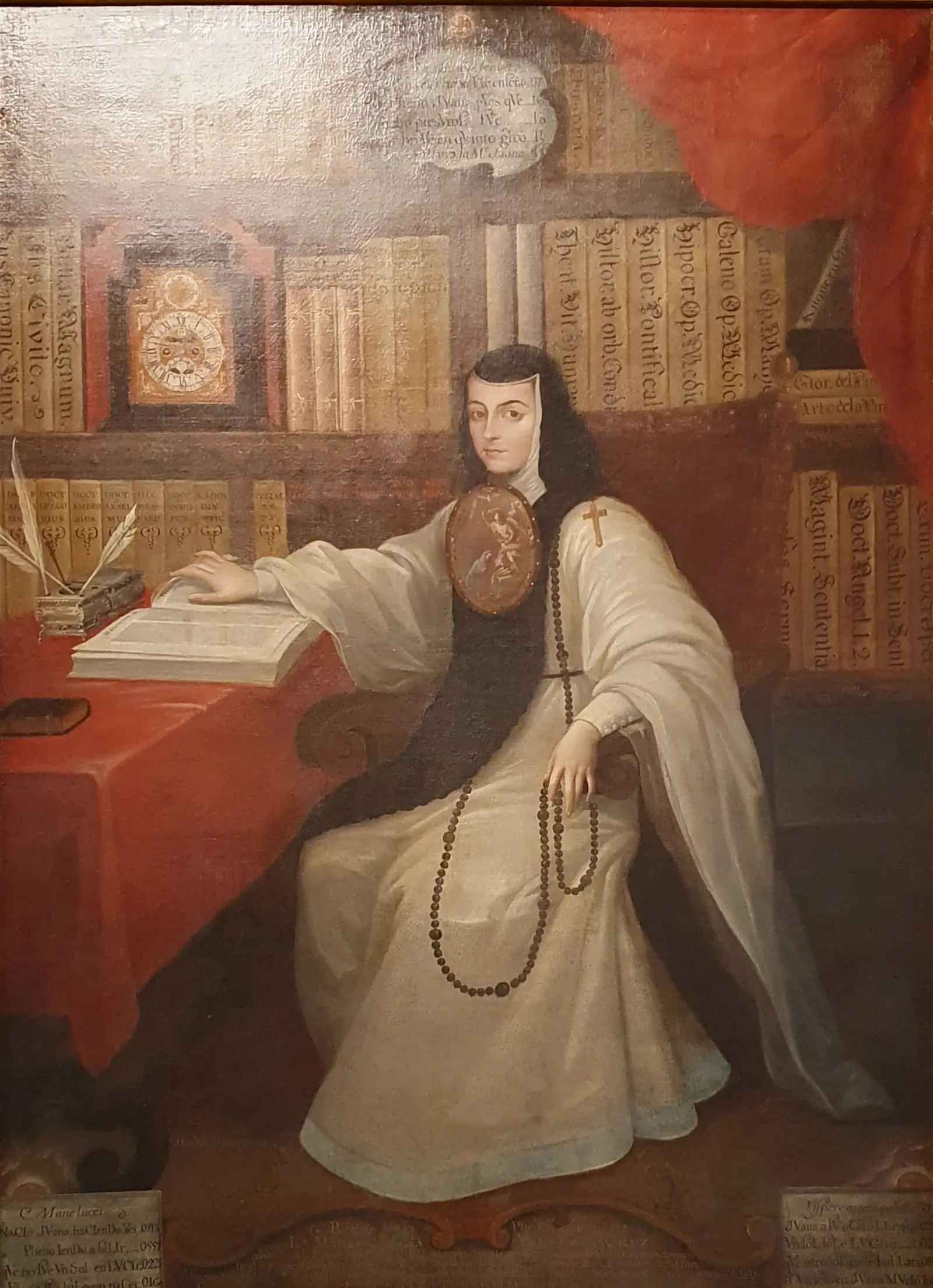 A nun in a white and black habit with a large painted badge on her chest sits in a library, one hand on an open book, with shelves of books behind her.