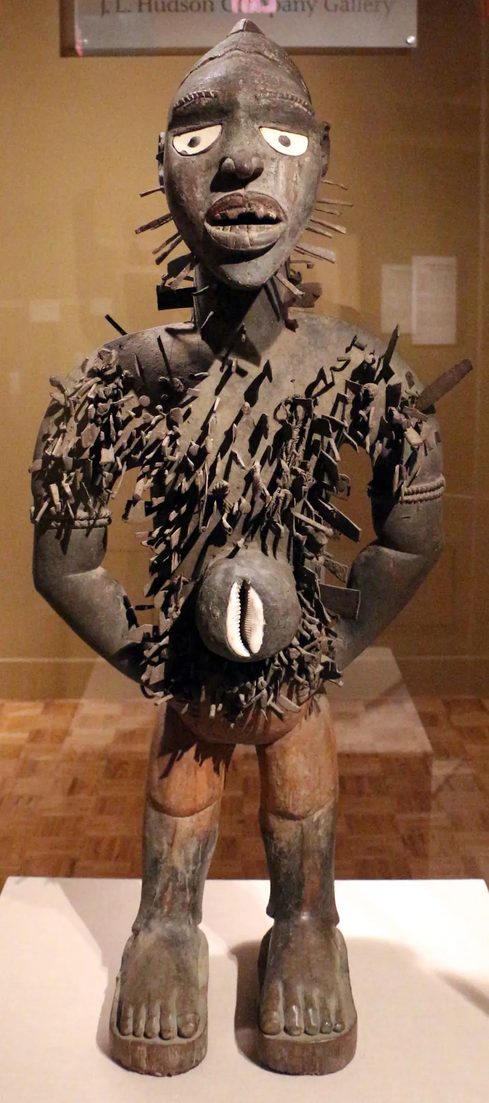A standing wooden figure with white inset eyes and hands on its hips, its chest and shoulders bristling with hundreds of nails and blades, and a shell set into its belly.