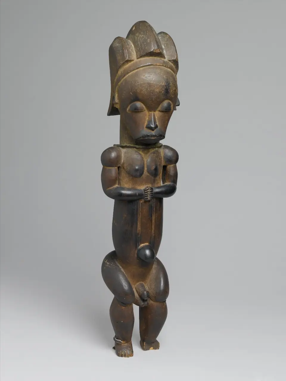 A dark, glossy wooden figure of a standing person with a high crested hairstyle, downcast eyes, hands held together at the chest and short, bent legs.