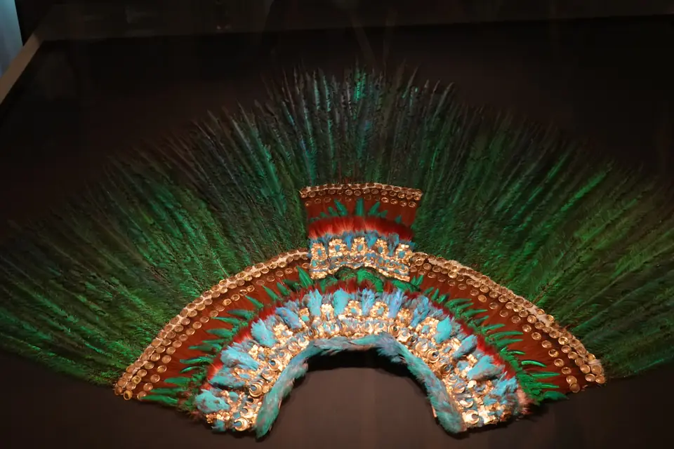 A large semicircular headdress of long green feathers with bands of blue feathers and small gold plates.