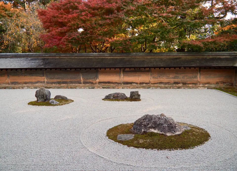A rectangular garden of raked white gravel with small groups of rocks surrounded by moss, behind a low wall.