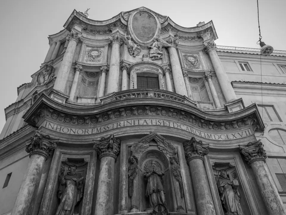 A narrow stone church facade whose surface curves in and out, with columns, niches with statues and an oval medallion at the top.