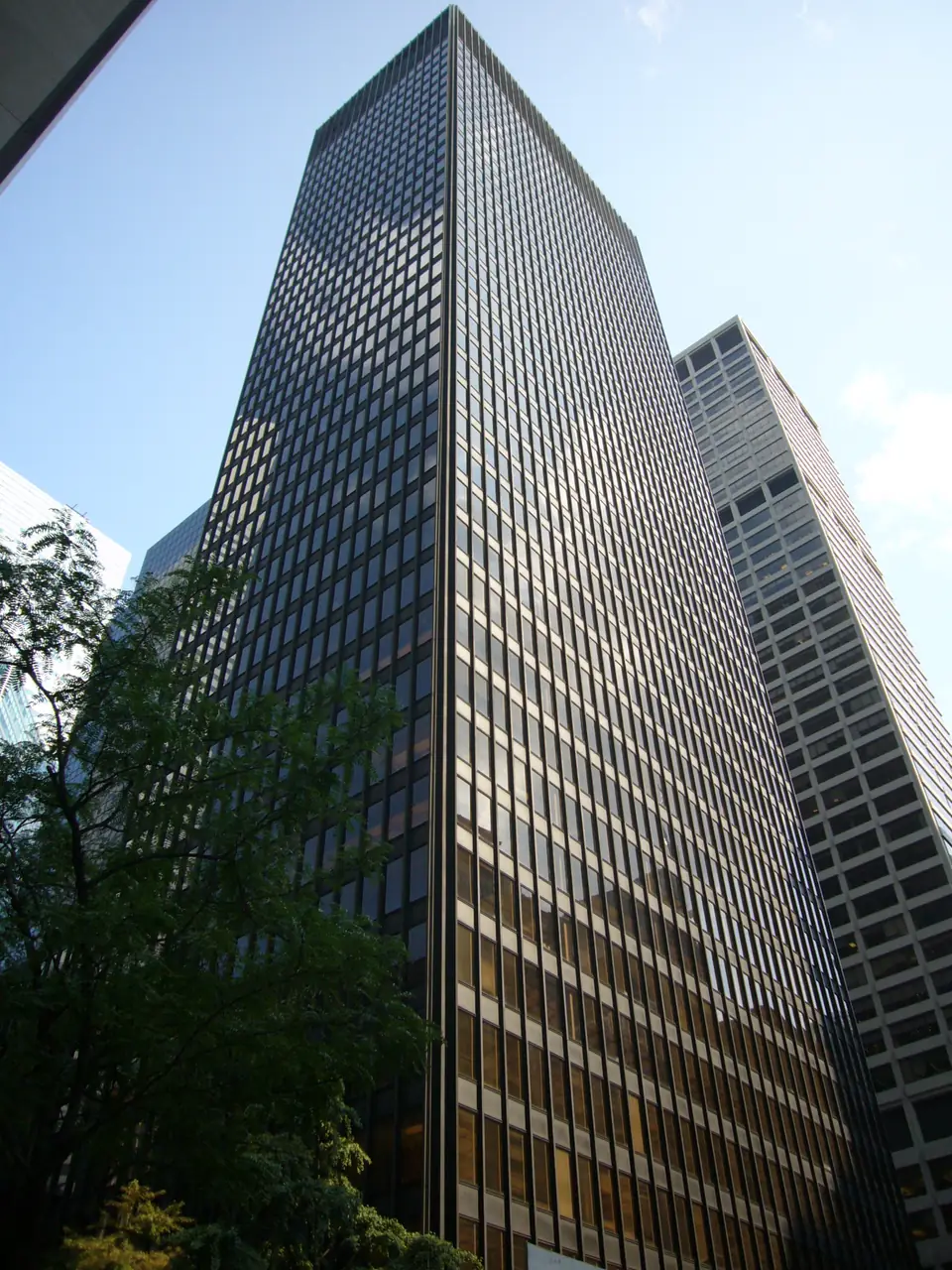A tall, dark tower of glass and bronze-colored metal, seen from below beside trees.