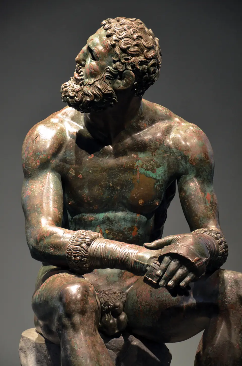 A bronze statue of a muscular seated man with a battered face, turning his head over his shoulder; his hands are wrapped in leather straps.