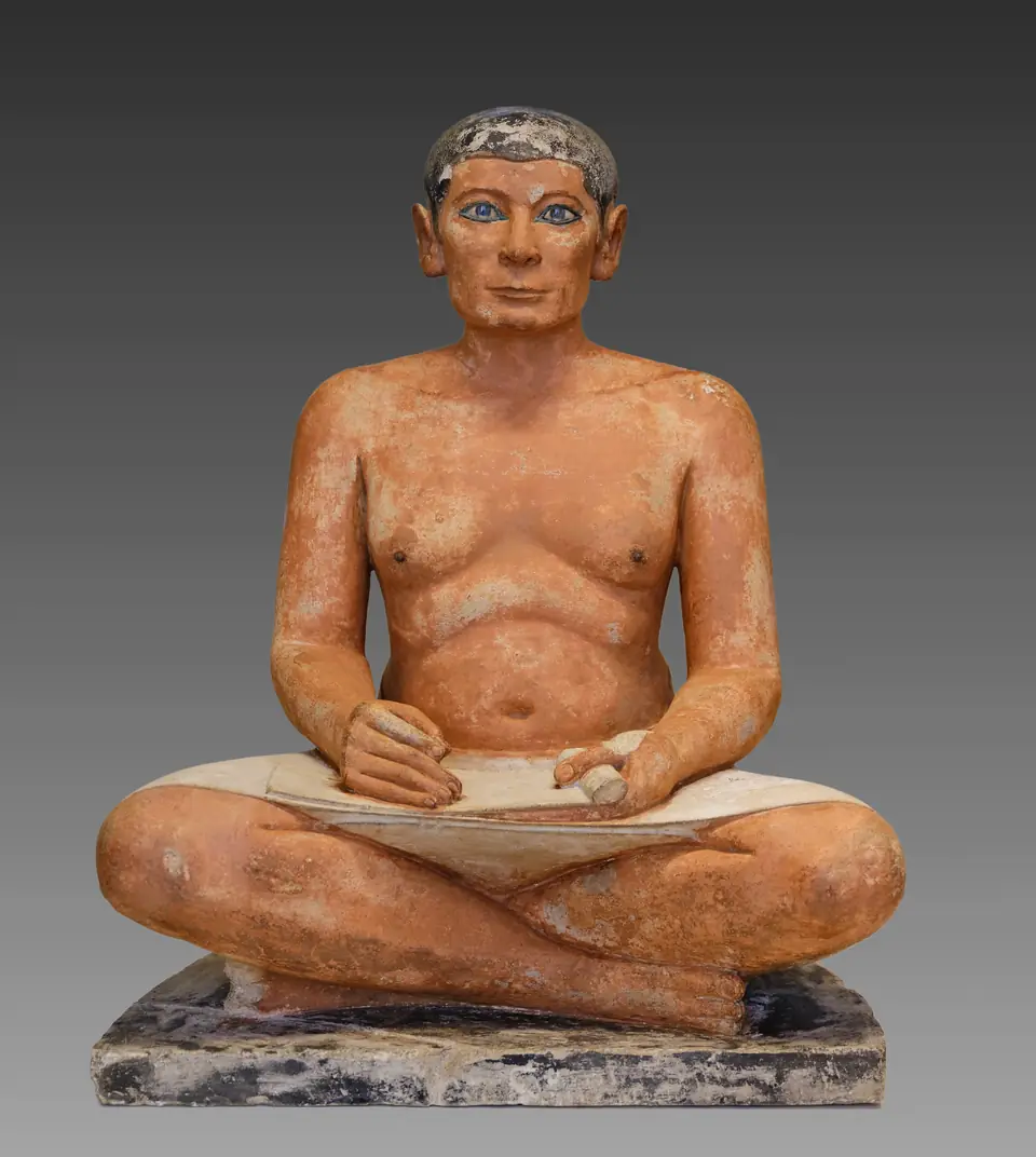 A painted stone statue of a seated man with crossed legs, a papyrus open on his lap and bright inlaid eyes.