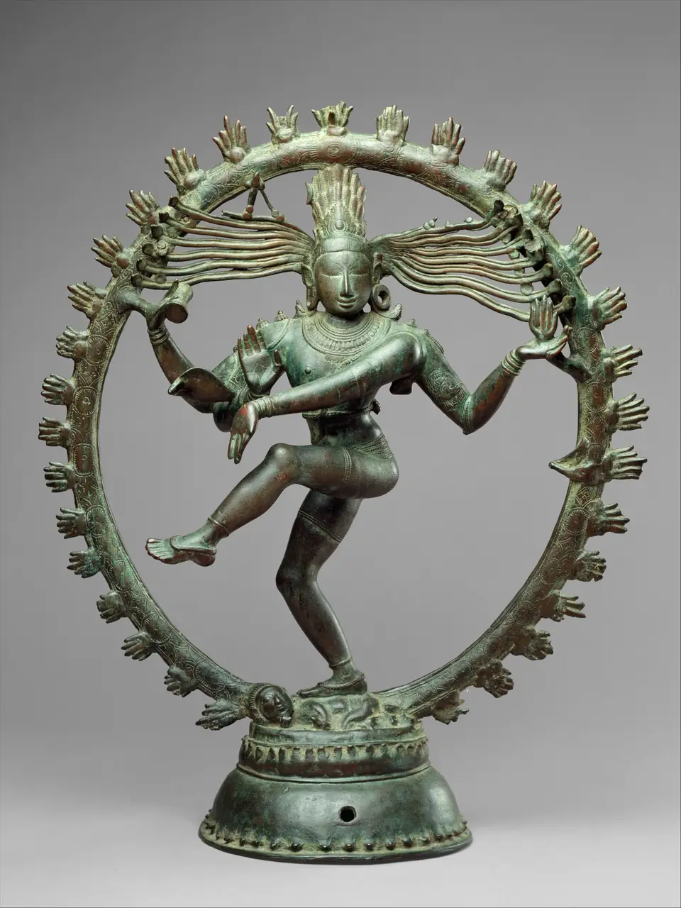A green-brown bronze statue of a four-armed dancing figure with one leg raised and hair streaming out to the sides, inside a ring of flames on a round lotus base.