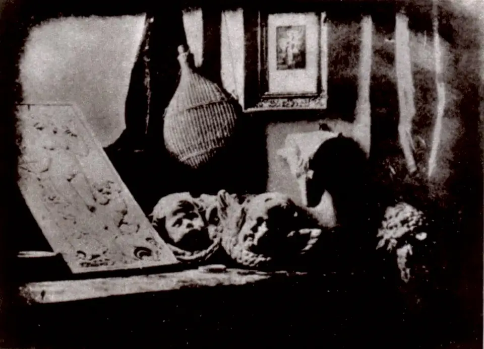 An early photograph of plaster casts, a wicker-covered bottle and a framed picture arranged on a shelf.