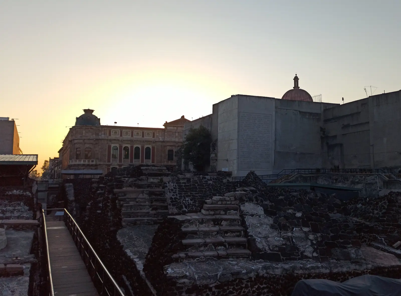 Excavated stone walls and stairways from a temple's successive building stages, surrounded by a modern city.
