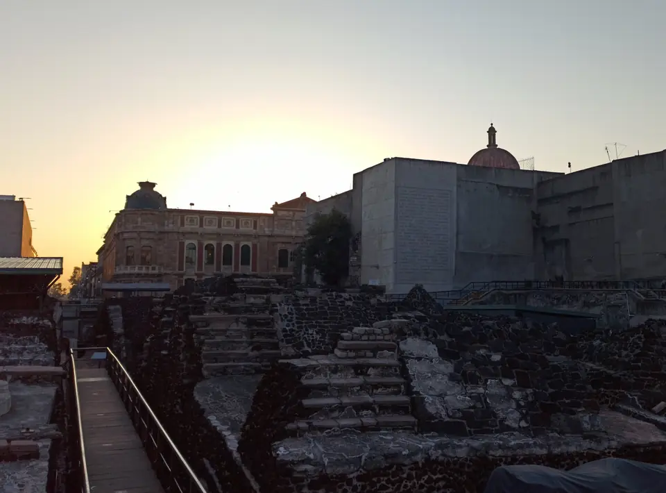 Excavated stone walls and stairways from a temple's successive building stages, surrounded by a modern city.