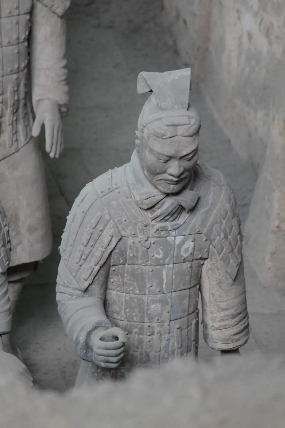 A life-size clay soldier in armor with a carved face and topknot, half-buried in earth in an excavation pit beside other figures.