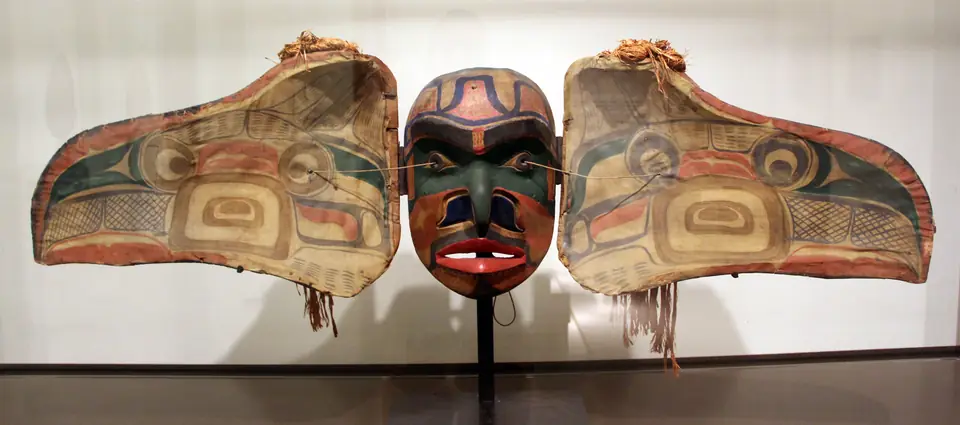 A carved wooden mask spread open into two large curved side panels painted with eyes and swirling shapes in red, green and black, flanking a painted human face with red lips, with tufts of shredded bark on top.
