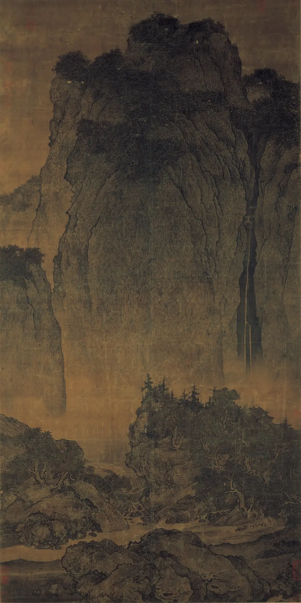 A tall hanging scroll of an enormous mountain towering over a waterfall, trees and a tiny mule train on a path below.