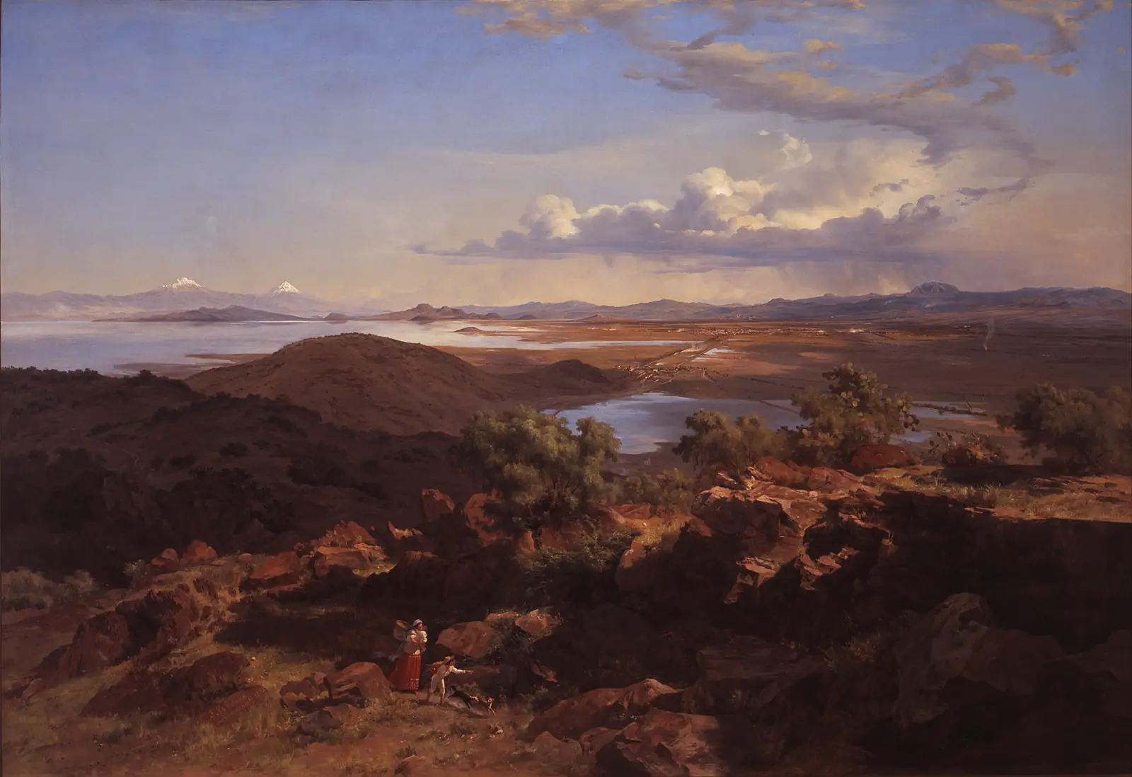 A wide, sunny panorama from a hillside over a valley with lakes, a distant city and snow-capped volcanoes.